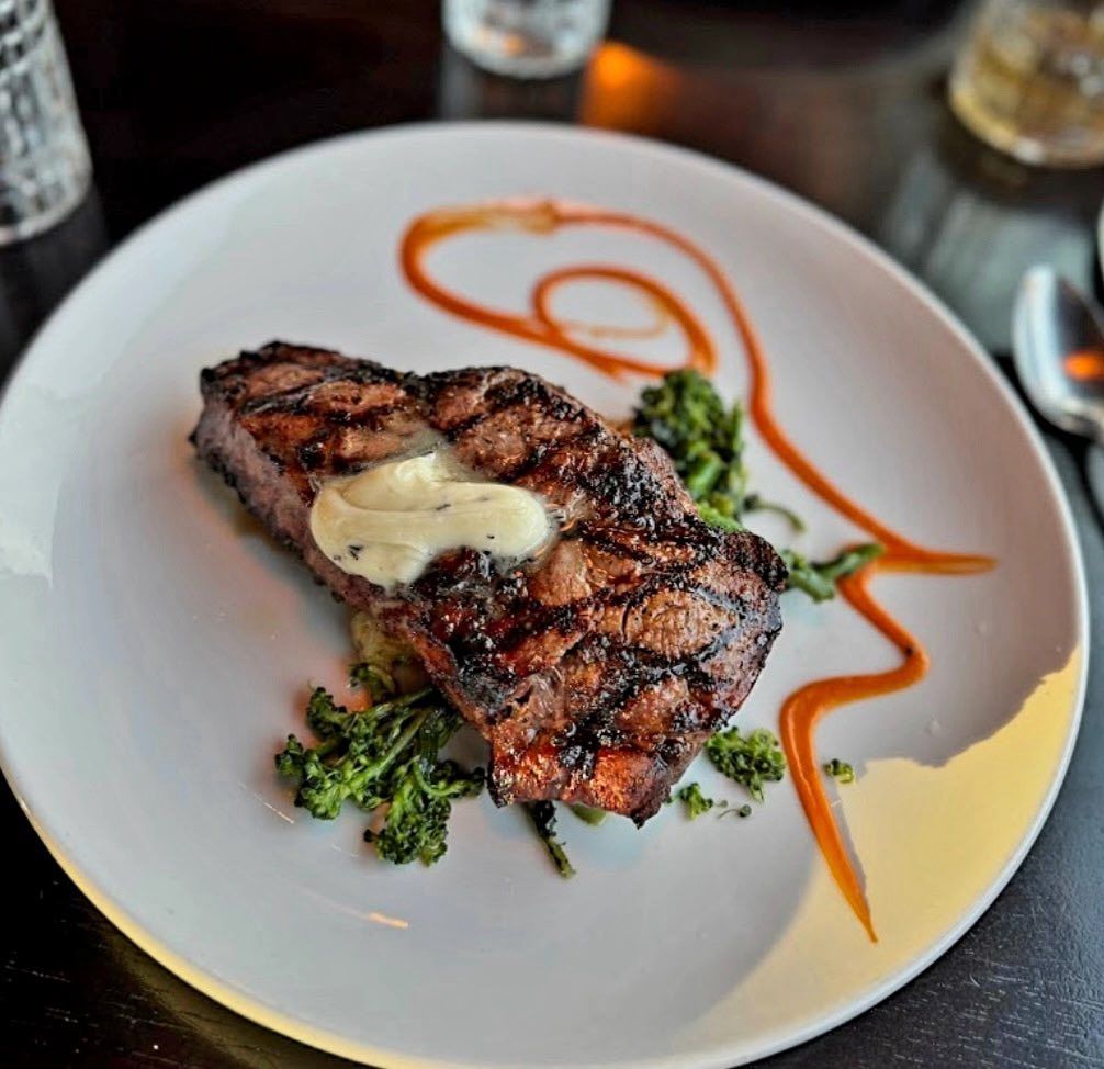 A grilled steak topped with melting butter sits on a white plate with greens and a decorative swirl of red sauce.