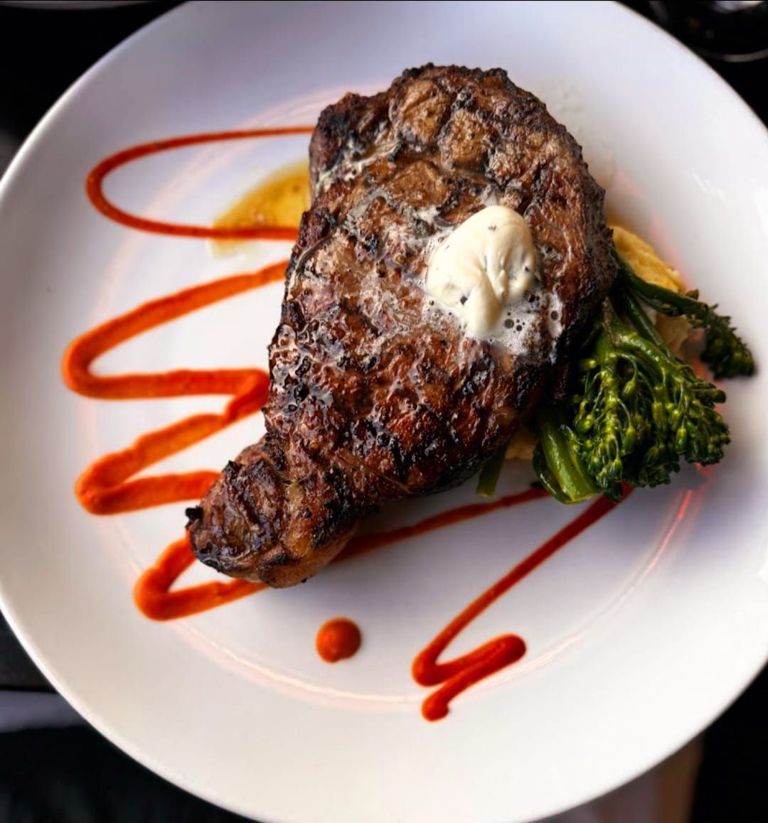 A seared steak topped with a pat of butter, served with broccolini on a plate decorated with zigzag red sauce.