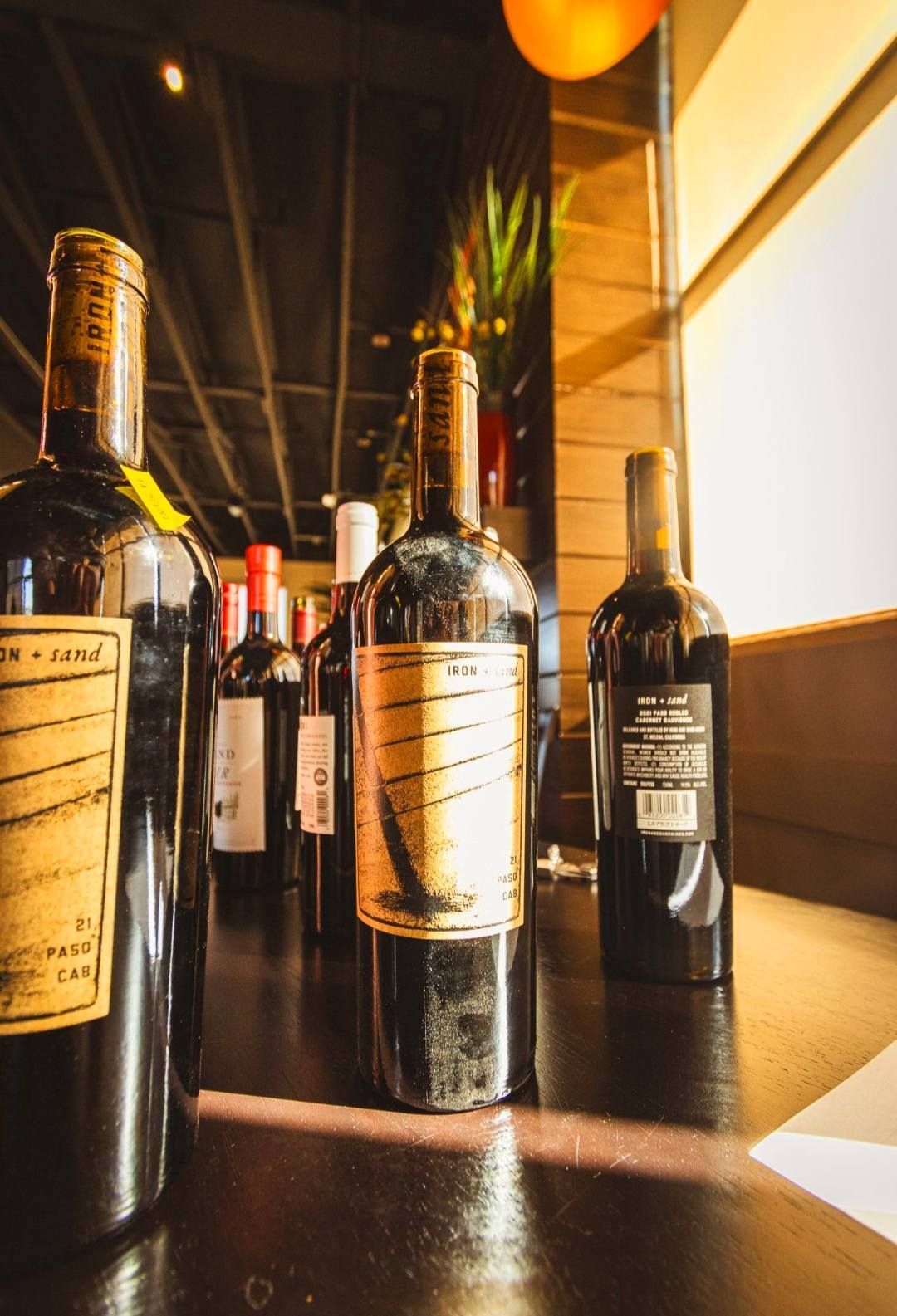 A row of dark wine bottles with tan labels sits on a reflective dark surface in a warmly lit room.