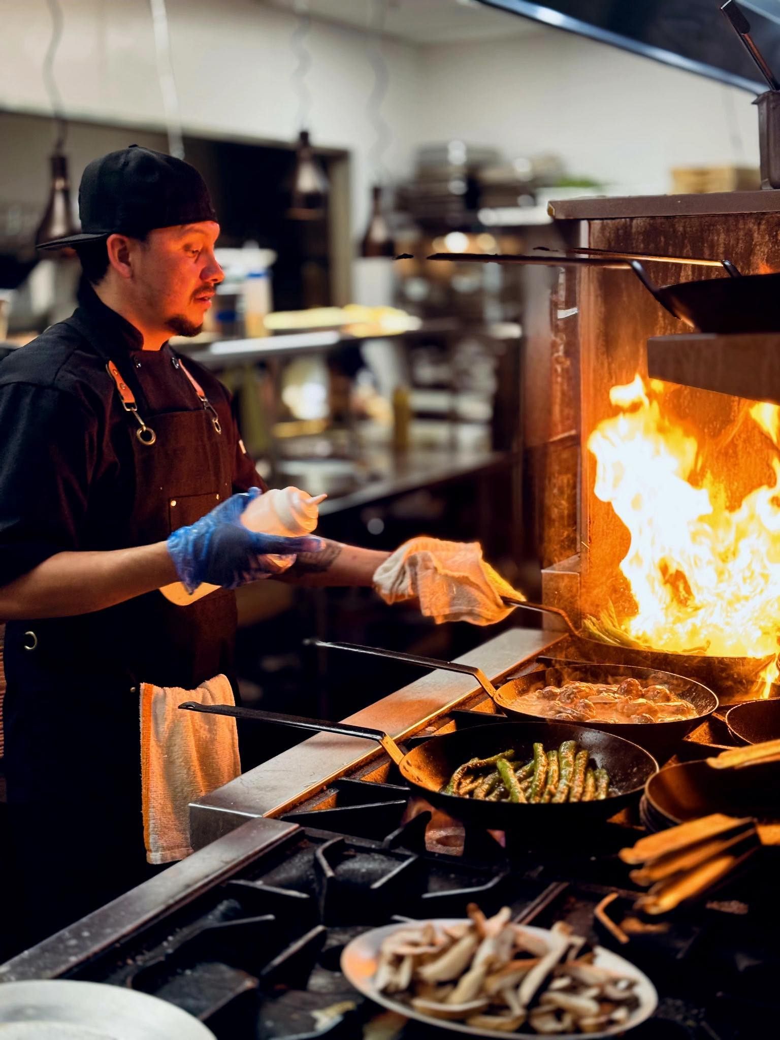 A chef in a professional kitchen uses a squeeze bottle while cooking with flames in pans on a stovetop.