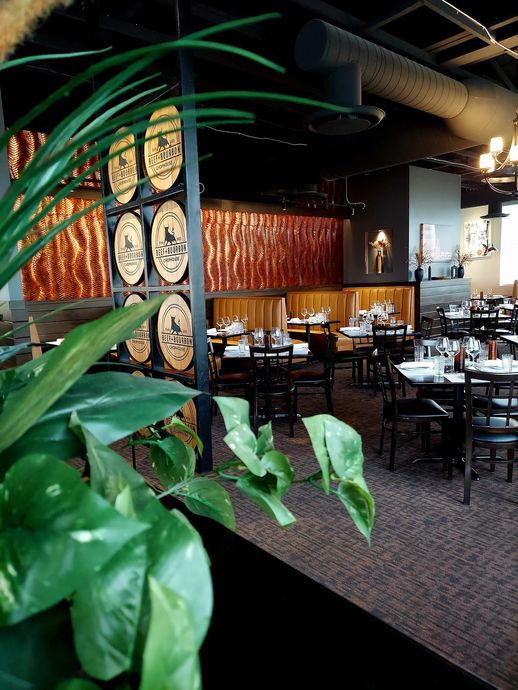 A dining area with round wood-cutout art on a metal partition, dark furniture, warm copper accents, and foreground plants.
