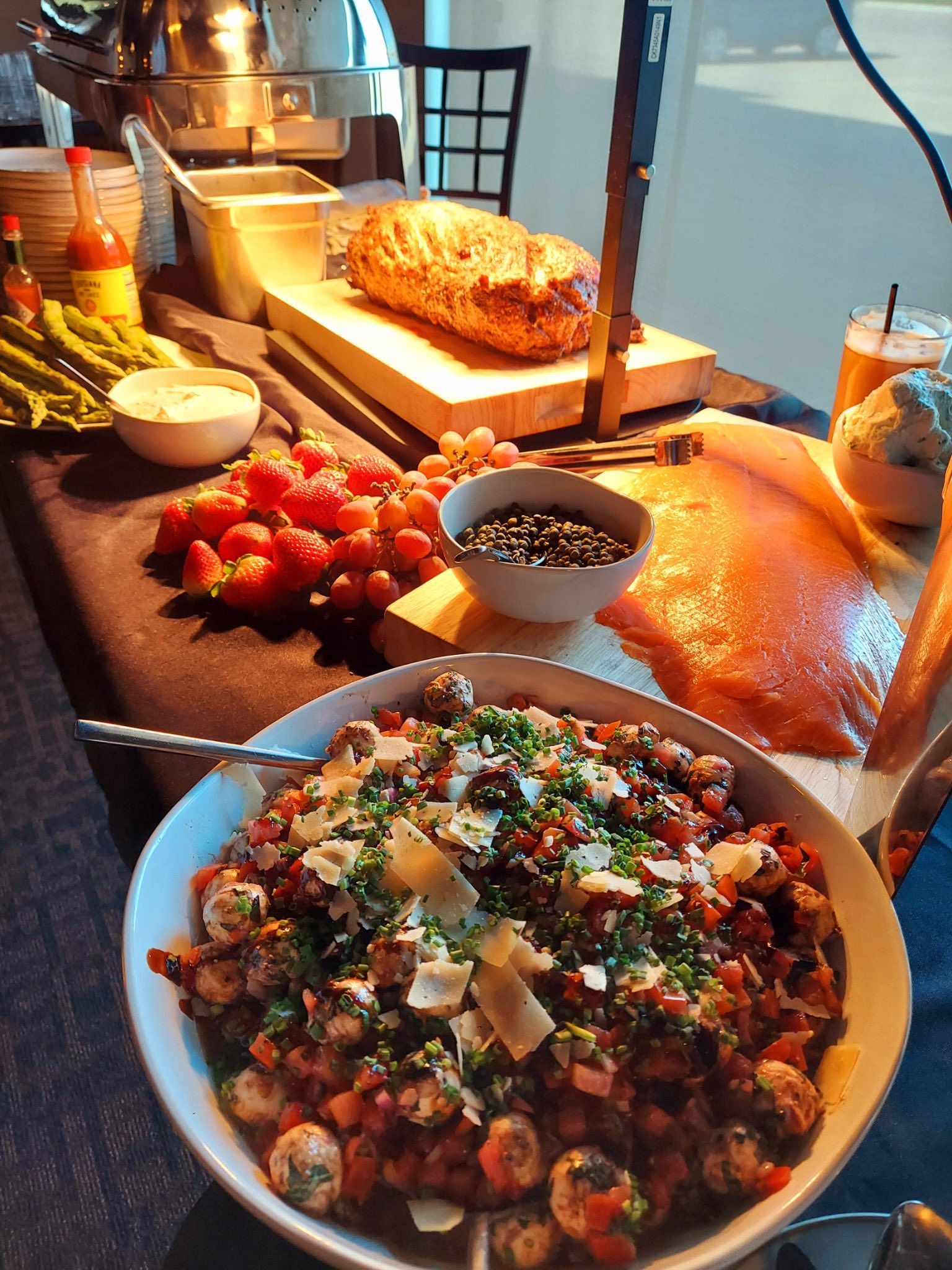 A buffet table featuring a large bowl of pasta with meatballs, sliced meat, fresh strawberries, and sides.