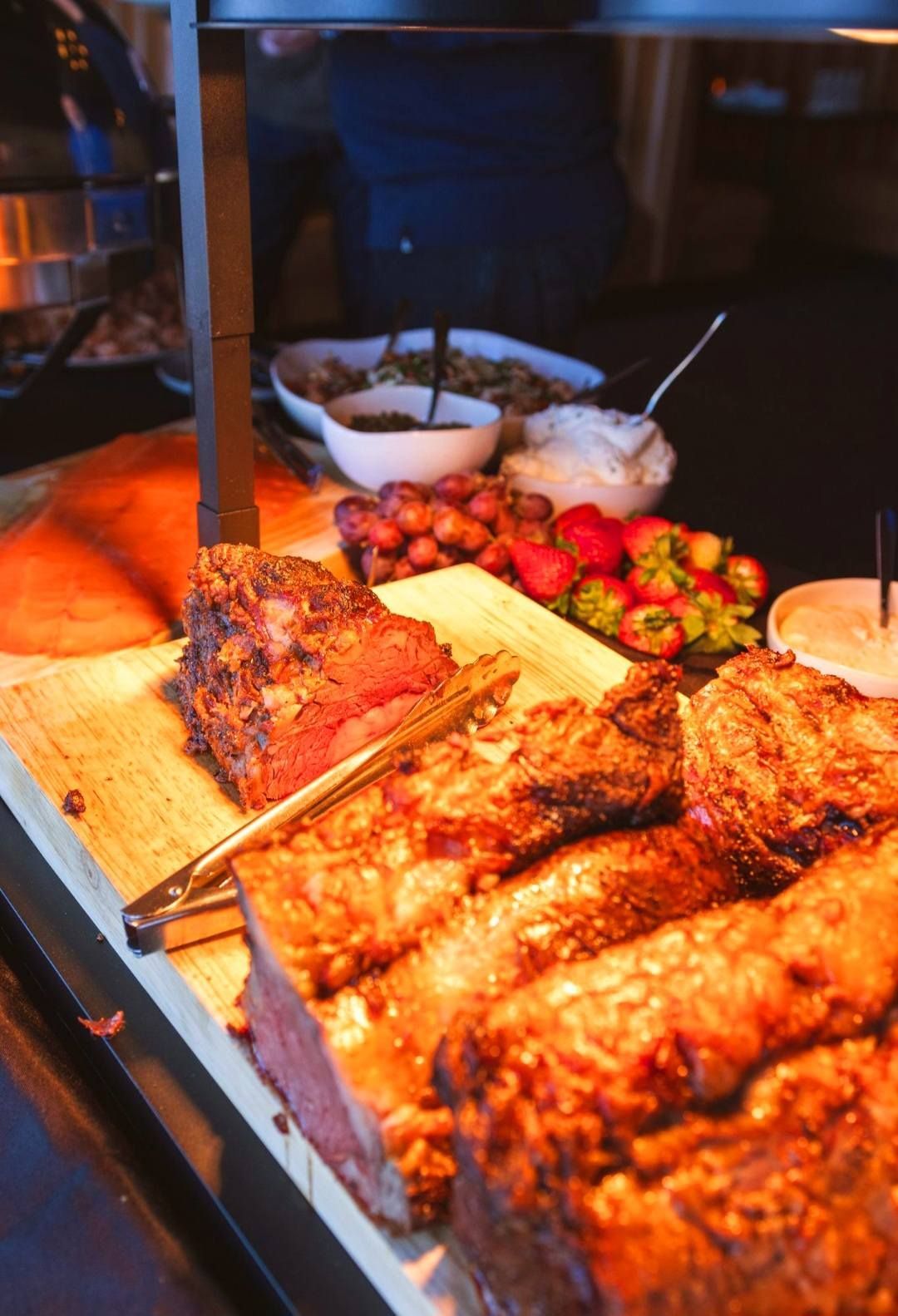 A buffet spread featuring carved roast beef, sliced salmon, grapes, strawberries, and bowls of accompaniments.