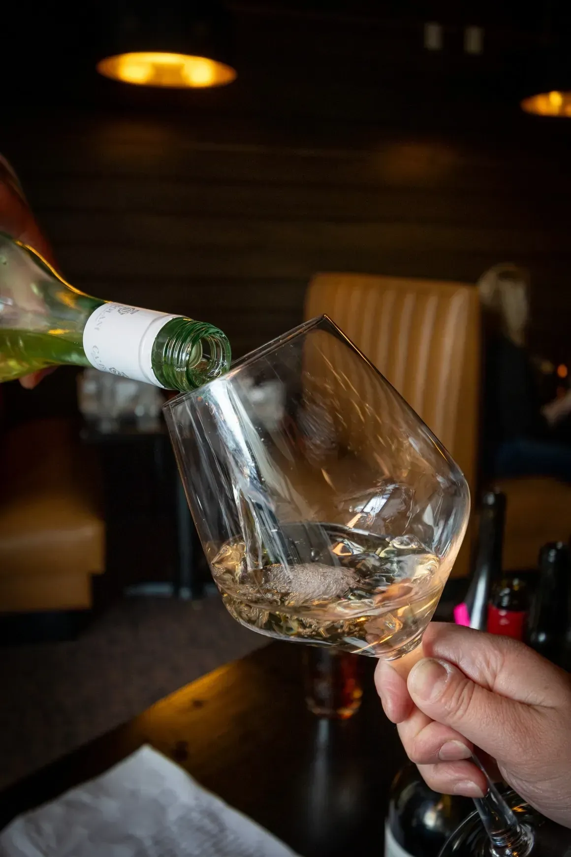 Wine being poured from a bottle into a clear glass held by a hand in a dimly lit restaurant.