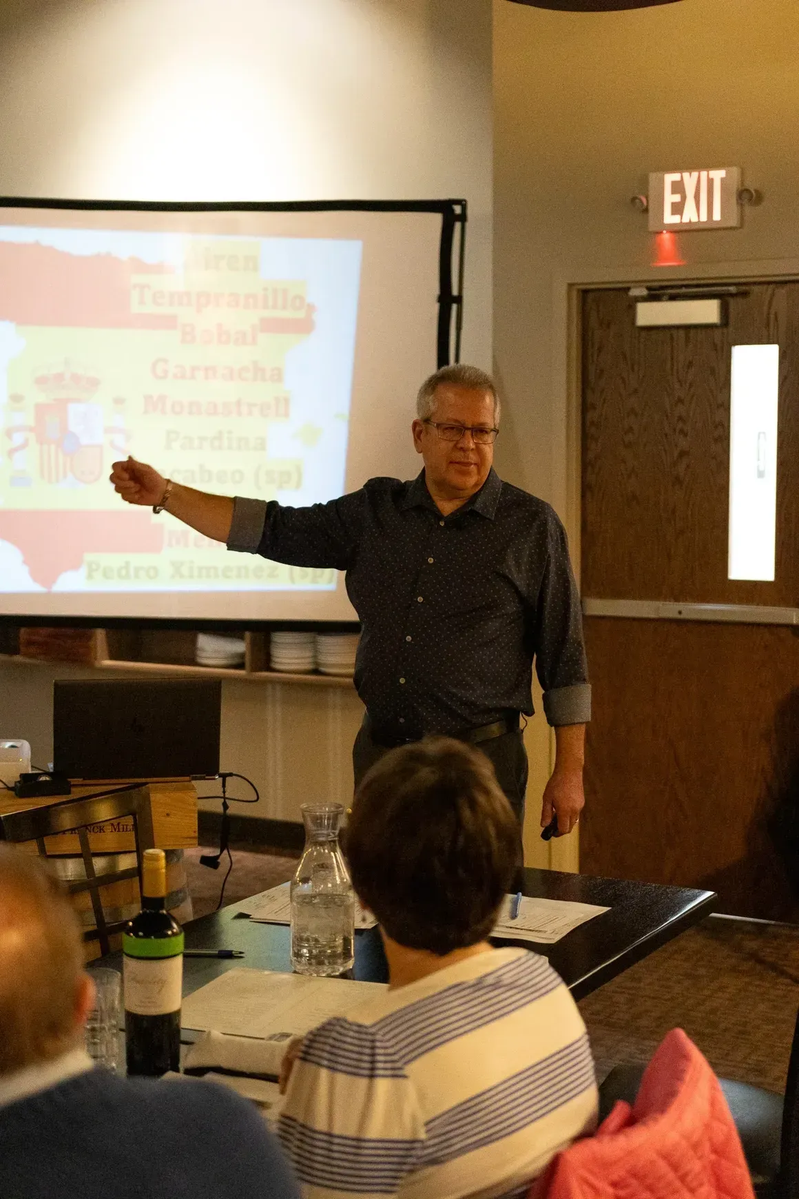 A speaker stands before a projector screen displaying text about Spanish wines, gesturing toward it for an audience.