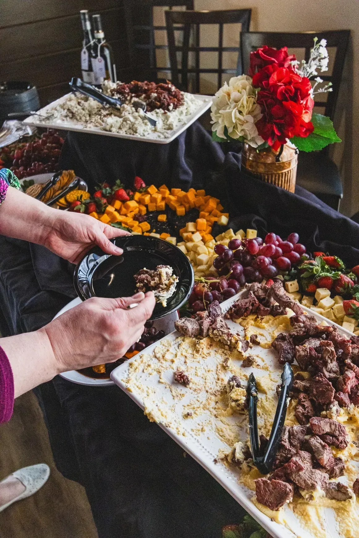 A person serves food onto a small black plate at a buffet table featuring meat, fruit, cheese, and a flower arrangement.