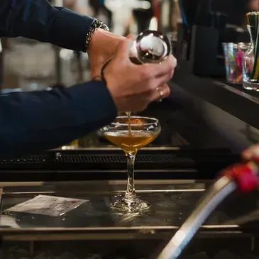 A person pours a golden cocktail from a metal shaker into a stemmed coupe glass at a dimly lit bar.