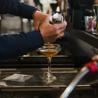 A person pours a golden cocktail from a metal shaker into a stemmed coupe glass at a dimly lit bar.