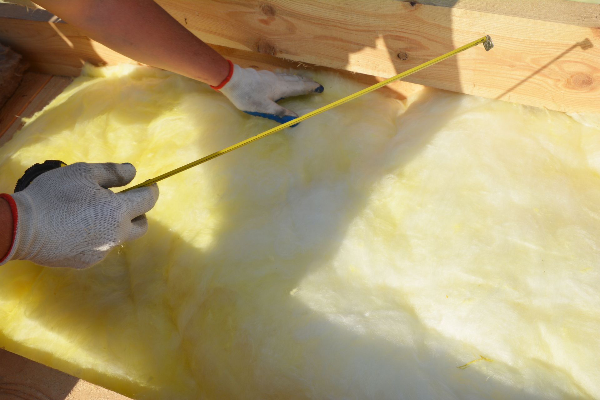 Gloved hands measuring yellow insulation with a tape measure in a wooden attic frame