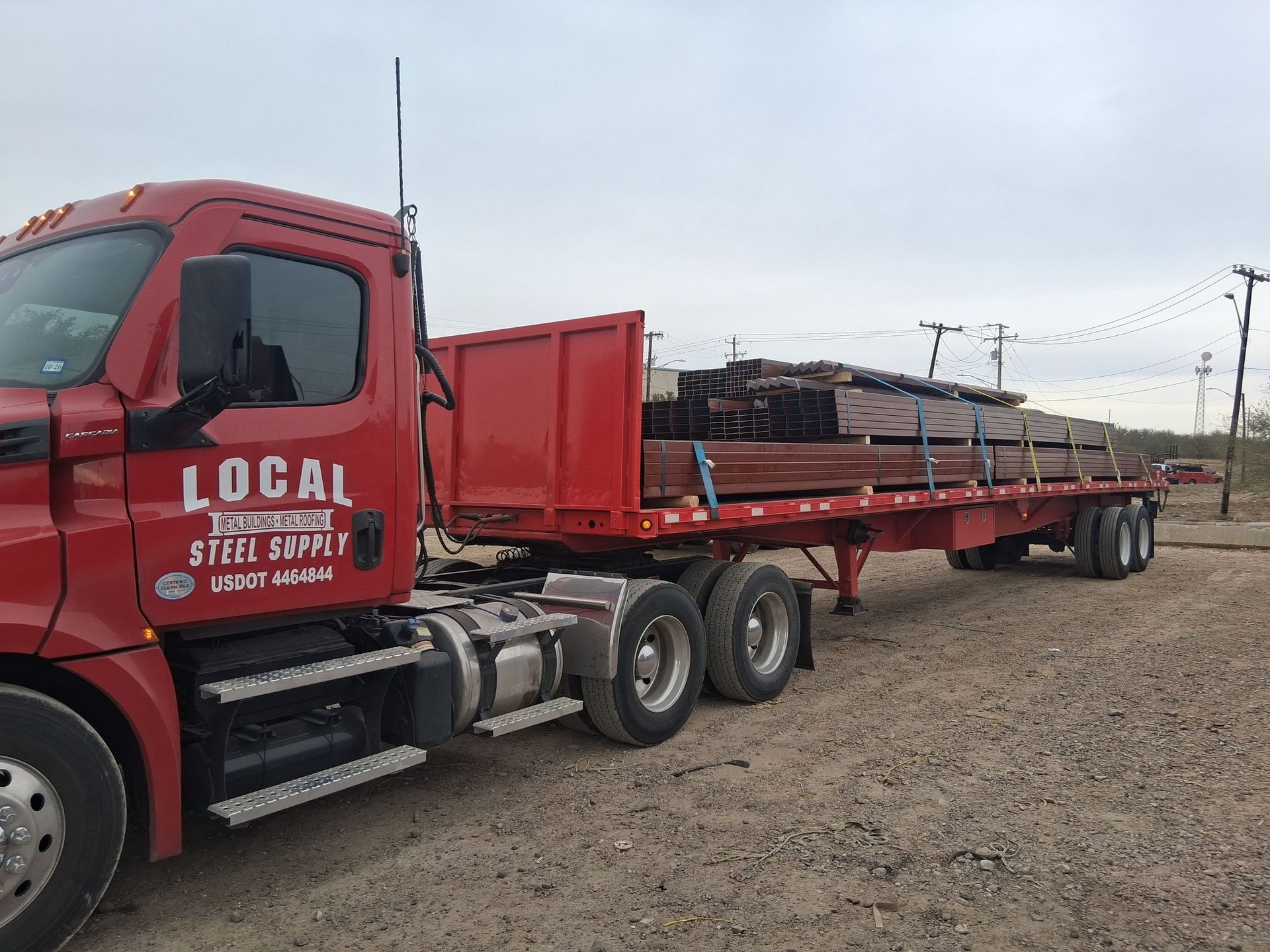 Red semi-truck hauling a long flatbed trailer loaded with lumber on a gravel lot.