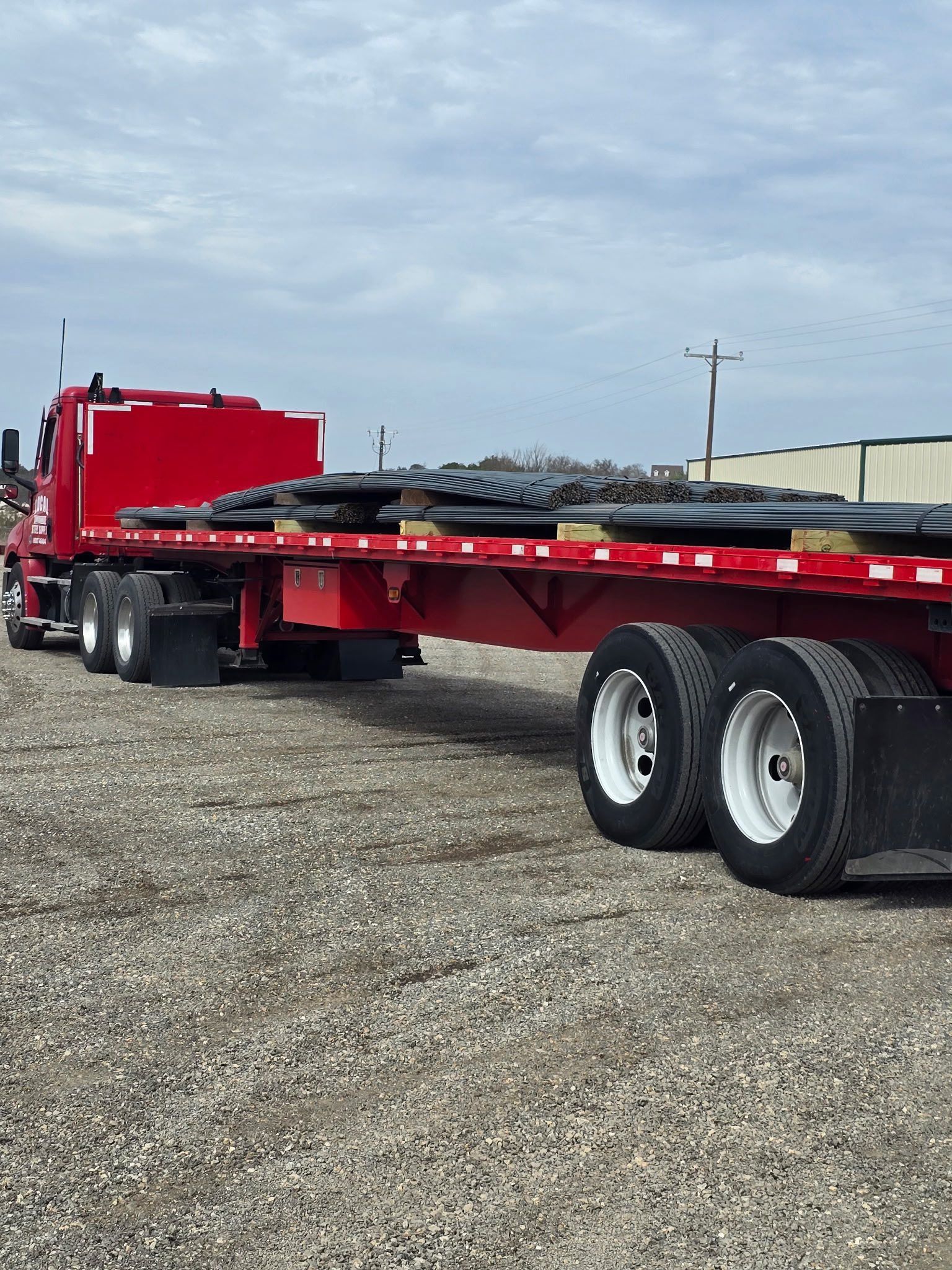 Red flatbed trailer truck parked on gravel under a cloudy sky.