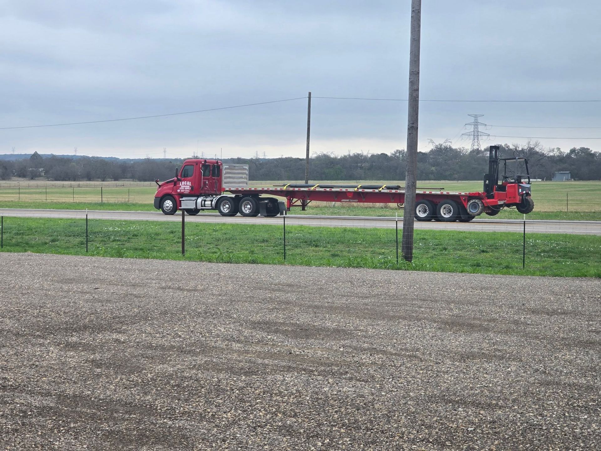 Red semi-truck hauling a long flatbed trailer across a grassy field under a cloudy sky.