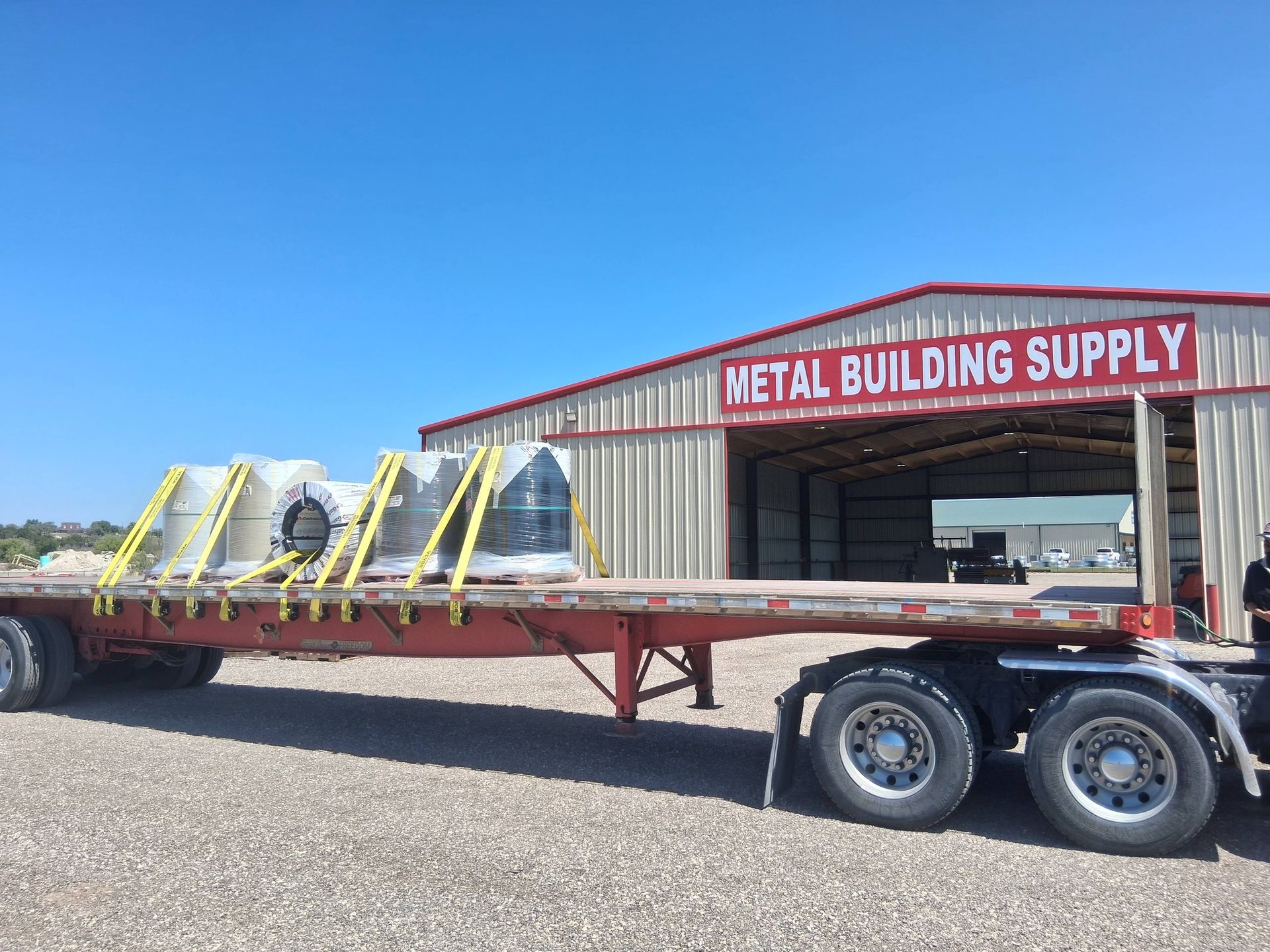 Flatbed trailer carrying metal tanks outside Metal Building Supply under a clear blue sky