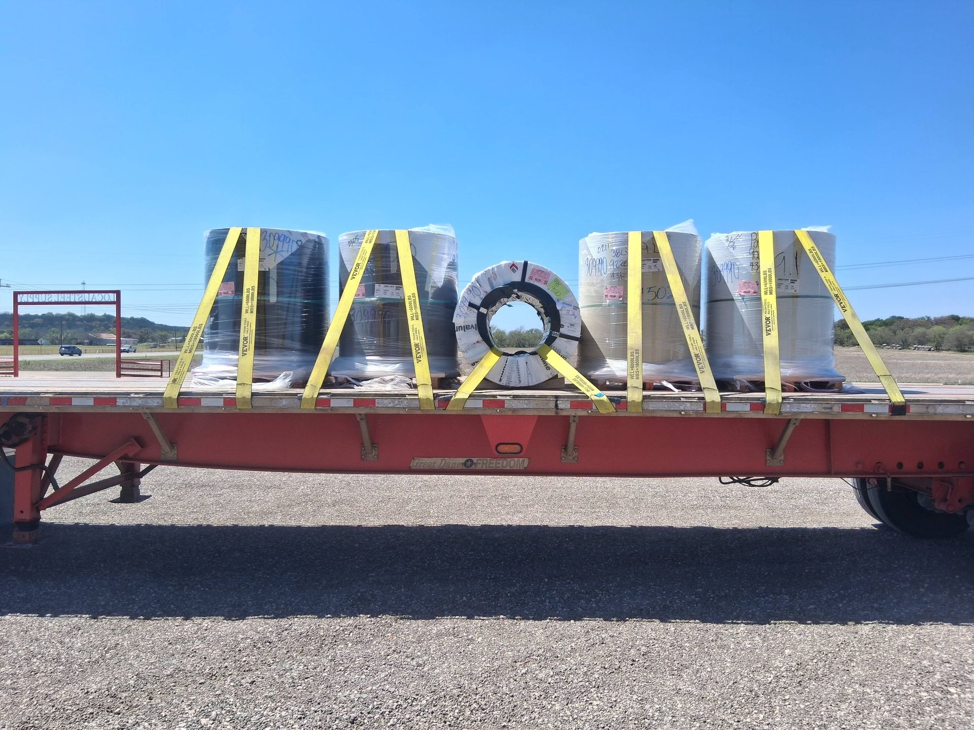 Red flatbed trailer carrying strapped industrial equipment outdoors on a gravel lot under a blue sky