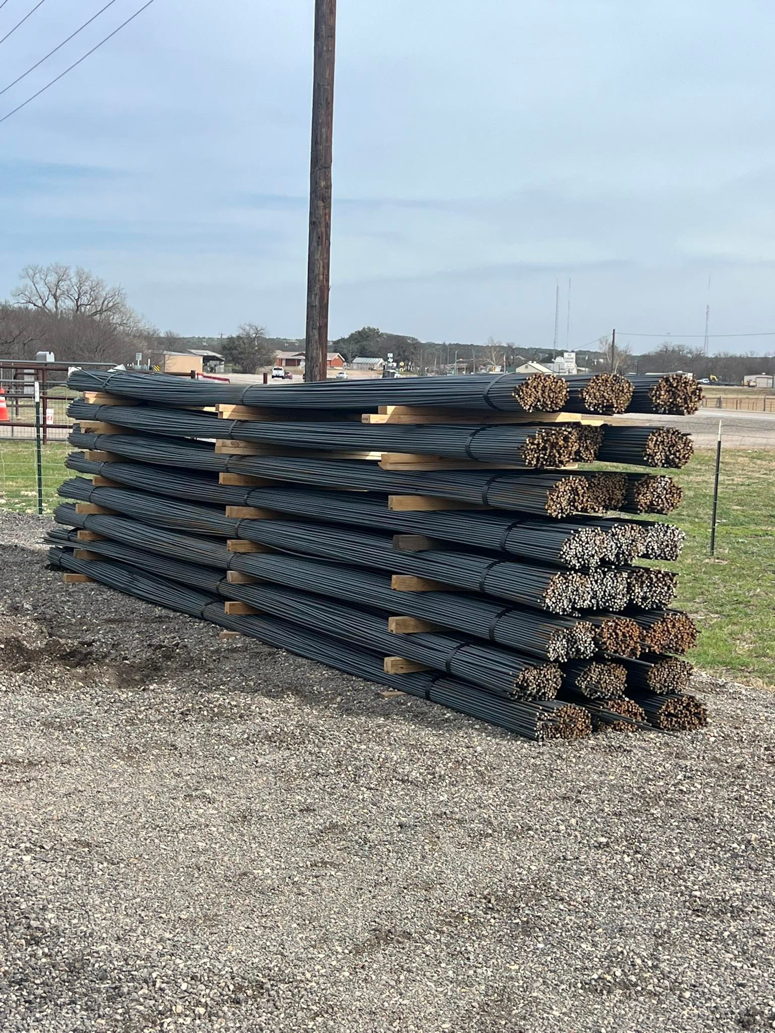 Stacked black metal fence panels on gravel in an outdoor yard, with a utility pole and sky in the background