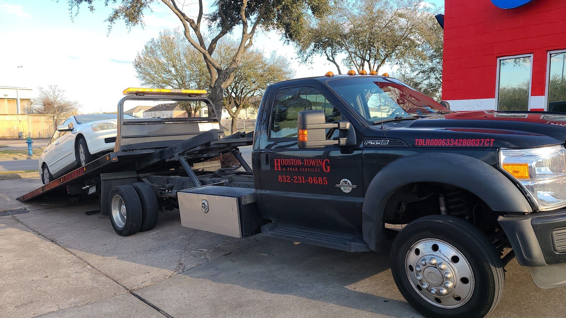 Tow truck carrying a black pickup on a flatbed in a driveway, with a red building and trees nearby | Rapid Repair Auto Center
