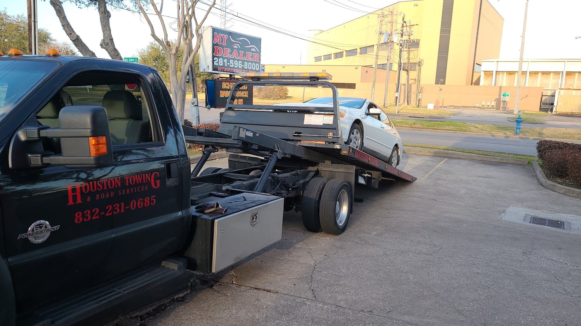 Tow truck hauling a black car with hazard lights on beside a roadside building. | Rapid Repair Auto Center