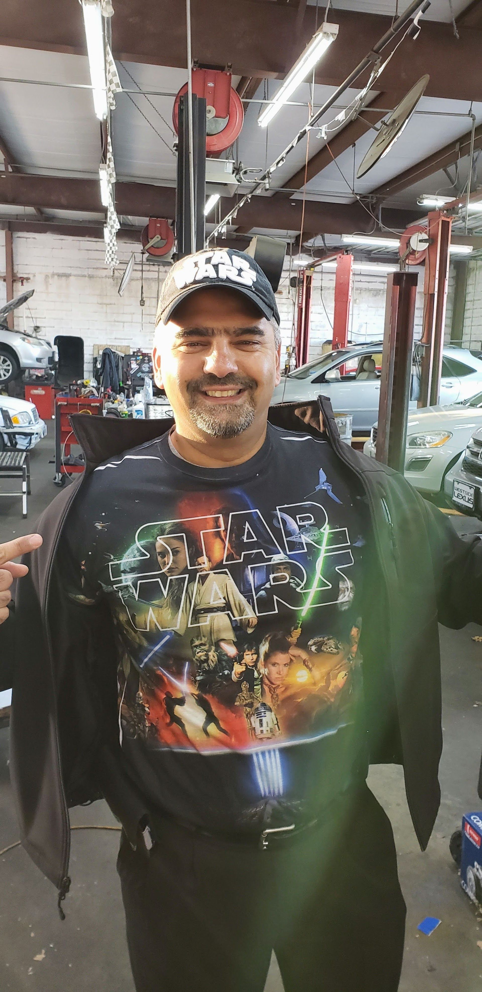 Smiling person in a cap and Star Wars shirt standing in a garage or auto shop. | Rapid Repair Auto Center