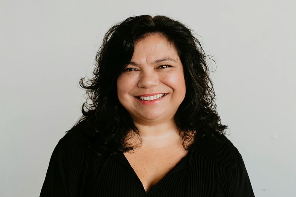 Smiling woman with curly dark hair wearing a black top against a light gray background