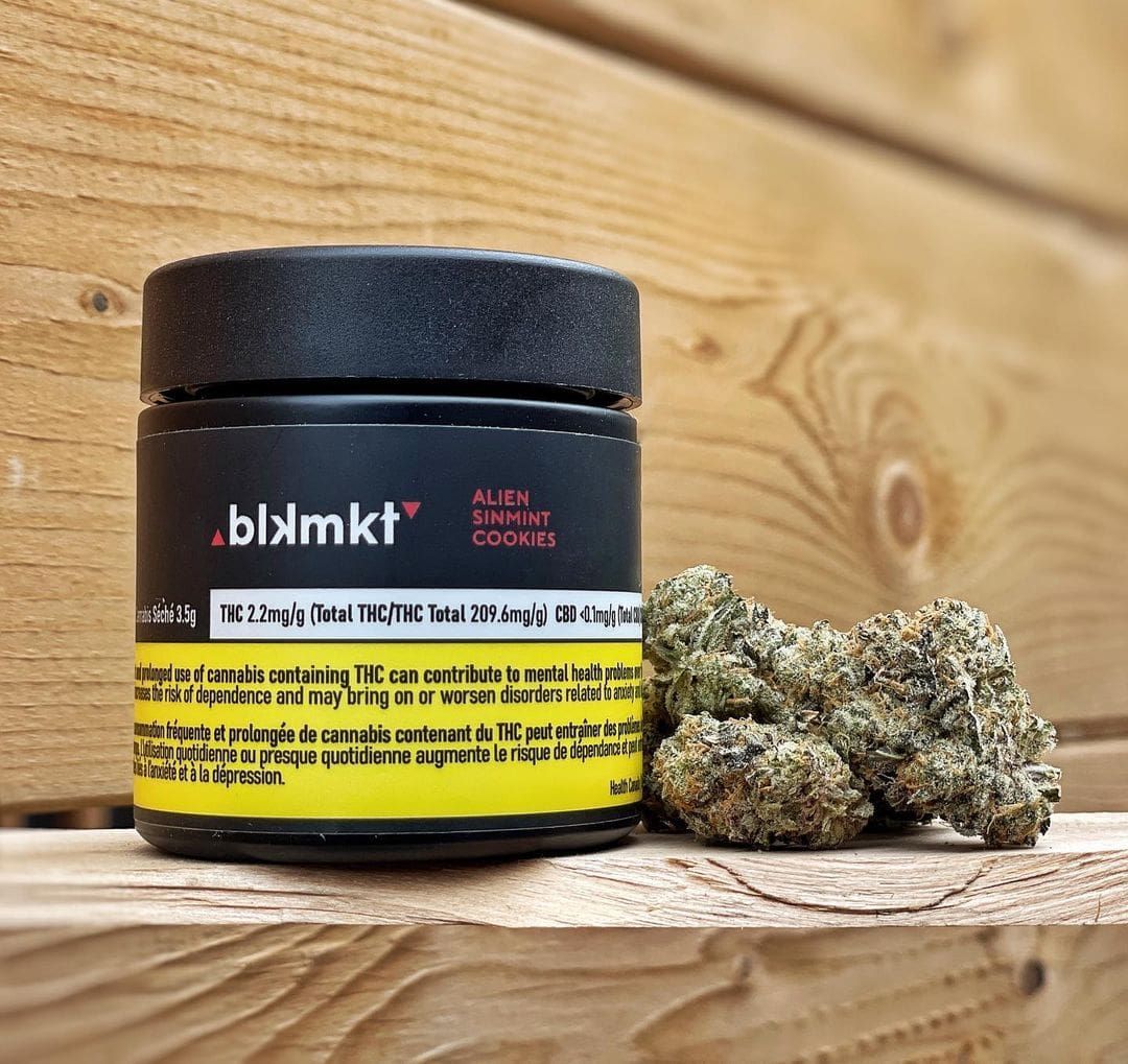 Black cannabis jar with yellow label beside cannabis buds on a wooden surface
