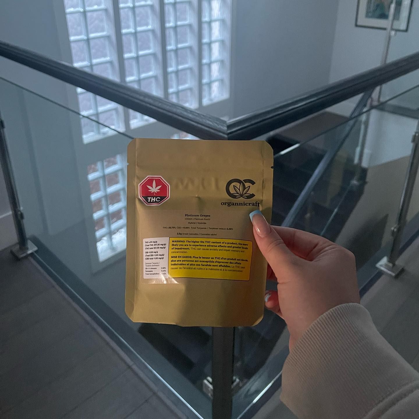 Hand holding a small brown package with labels beside a glass stairwell railing.