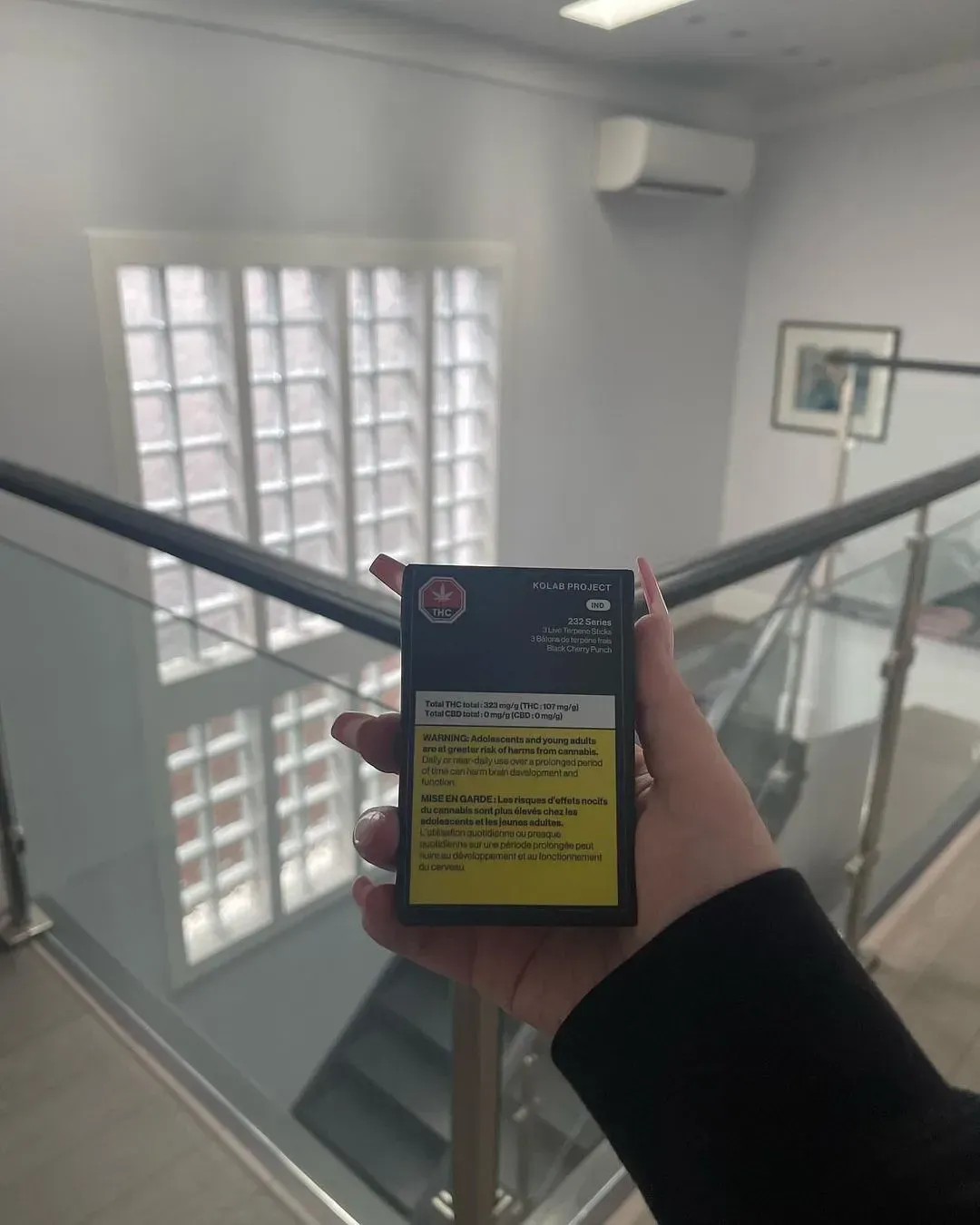 Hand holding a small black-and-yellow device on a stairwell landing with glass railing and windowed wall.