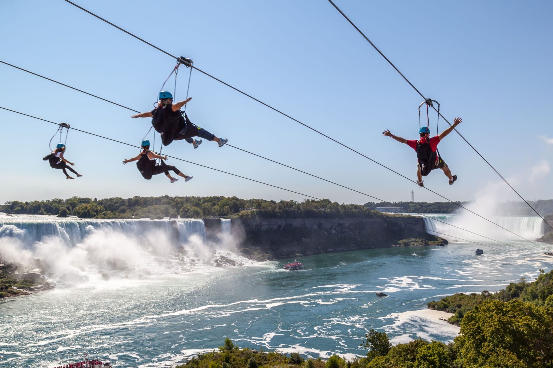 People zip lining over a wide waterfall and river with mist below on a sunny day