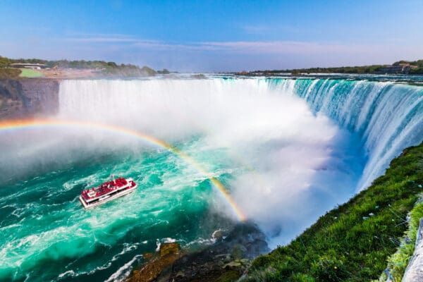 Tour boat beneath a rainbow at the base of a massive waterfall, with mist and turquoise water