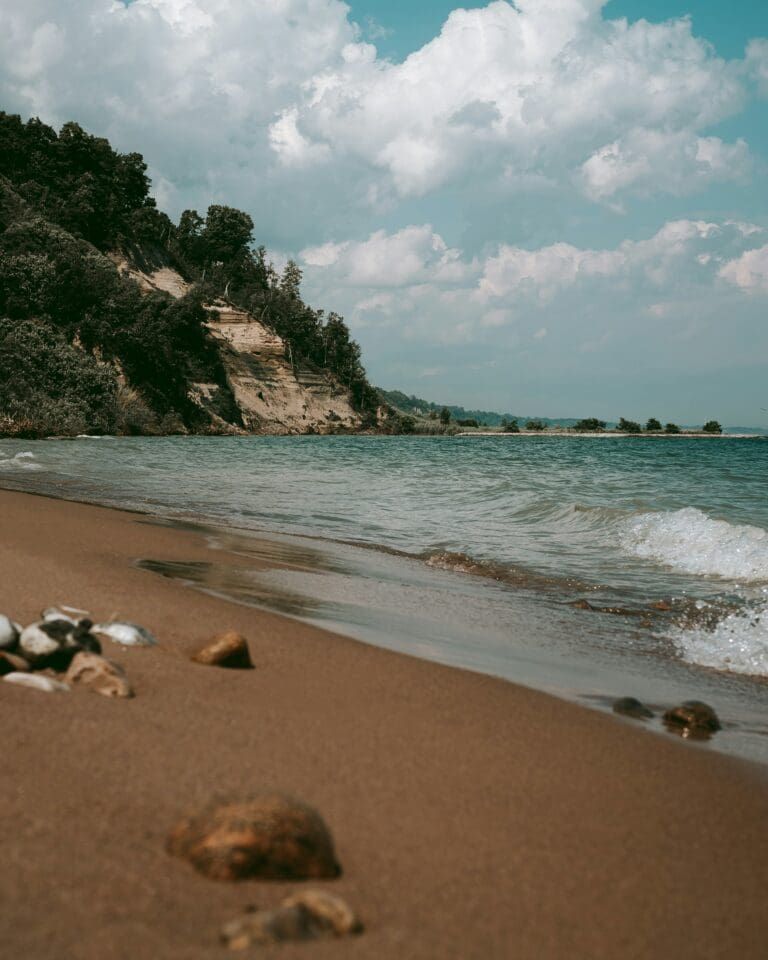 Sandy beach with scattered rocks, forested cliff, gentle waves, and cloudy sky