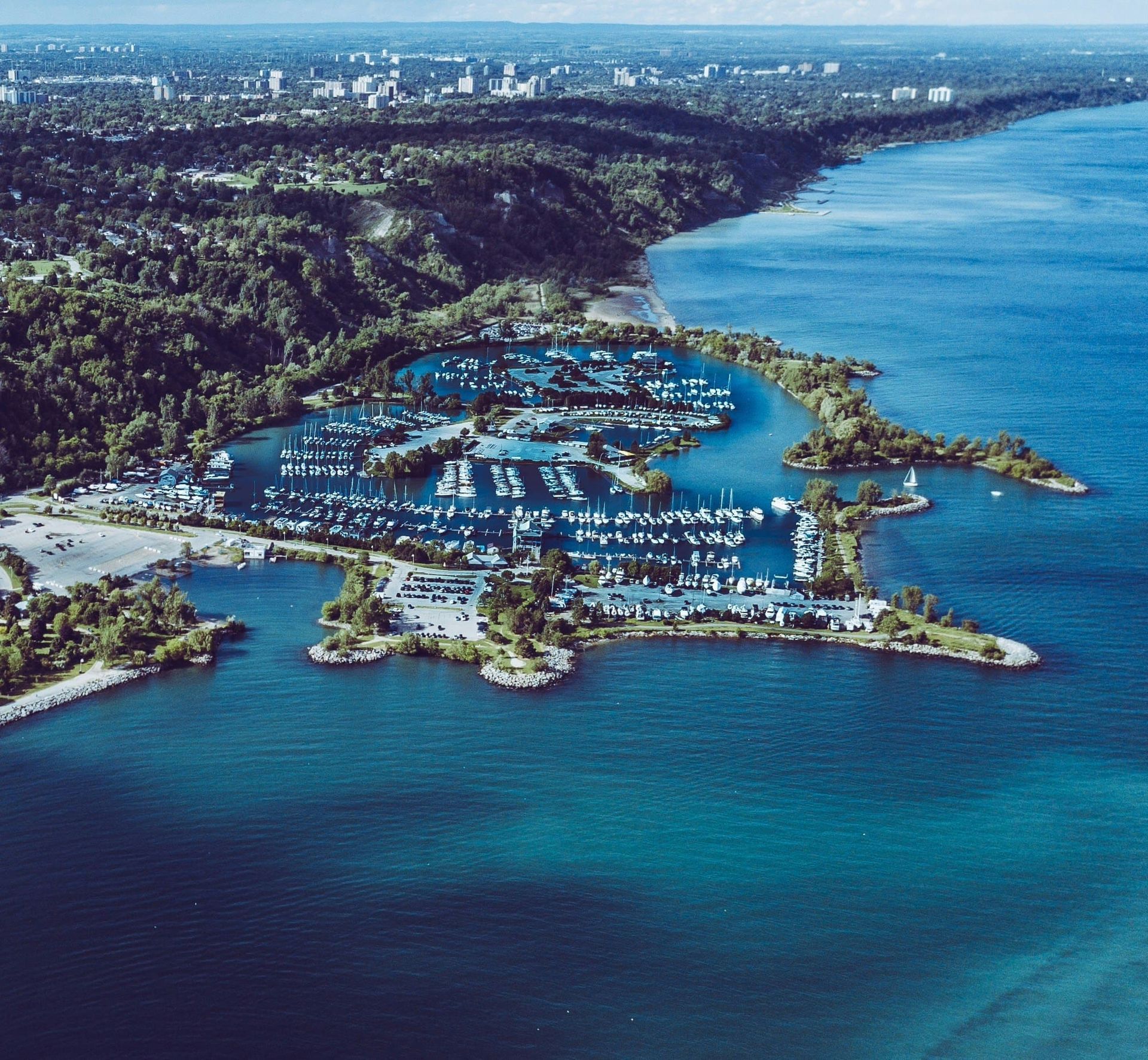 Aerial view of a coastal marina with docks, boats, and turquoise water beside a wooded shoreline.