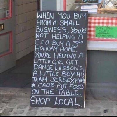 Handwritten chalkboard sign urging shoppers to buy local, with a red-checkered tablecloth beside it