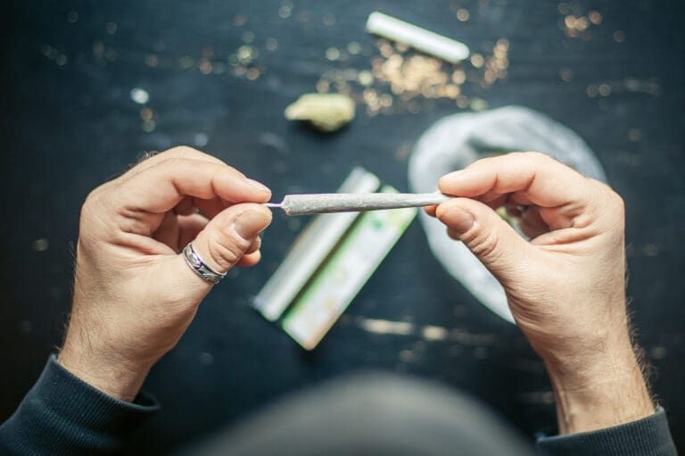 Hands holding a rolled joint over a dark table with cannabis and a pack of rolling papers.