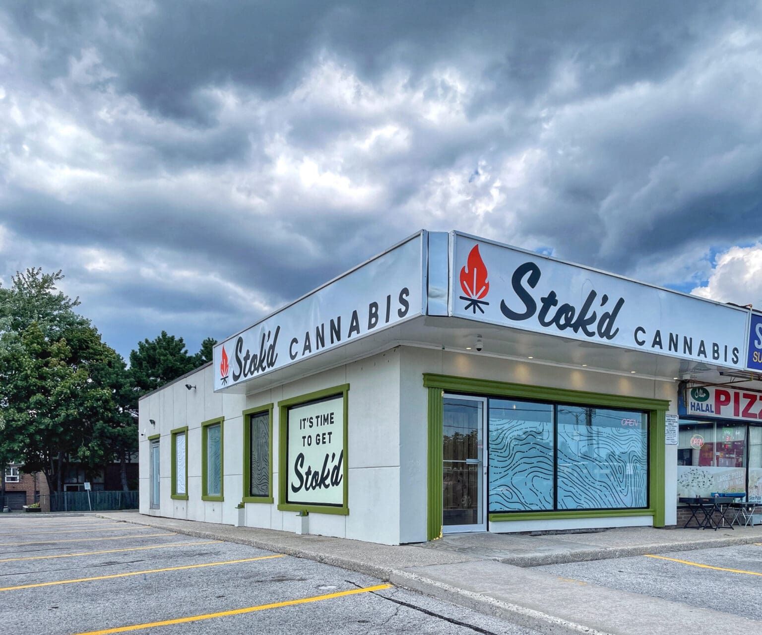Corner cannabis shop with white-and-green signage under stormy clouds, empty parking lot in front.