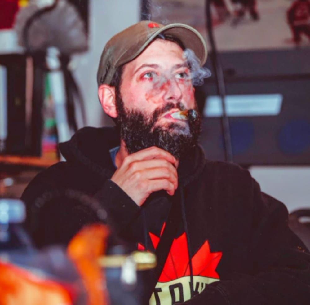 Bearded man in a cap and black hoodie, seated indoors and looking upward with a thoughtful expression