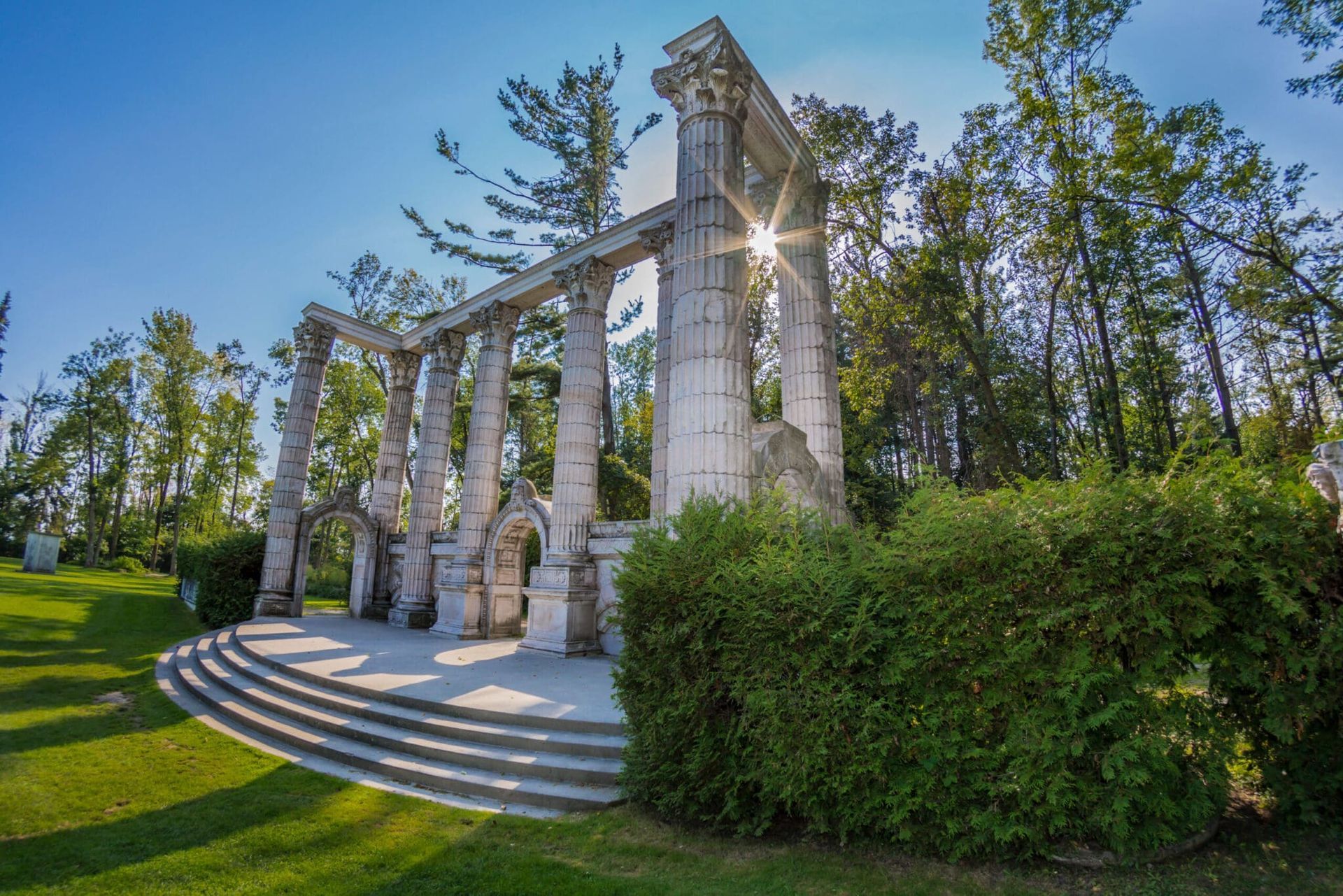 Stone memorial pavilion with columns and steps in a sunny park, surrounded by trees and bushes