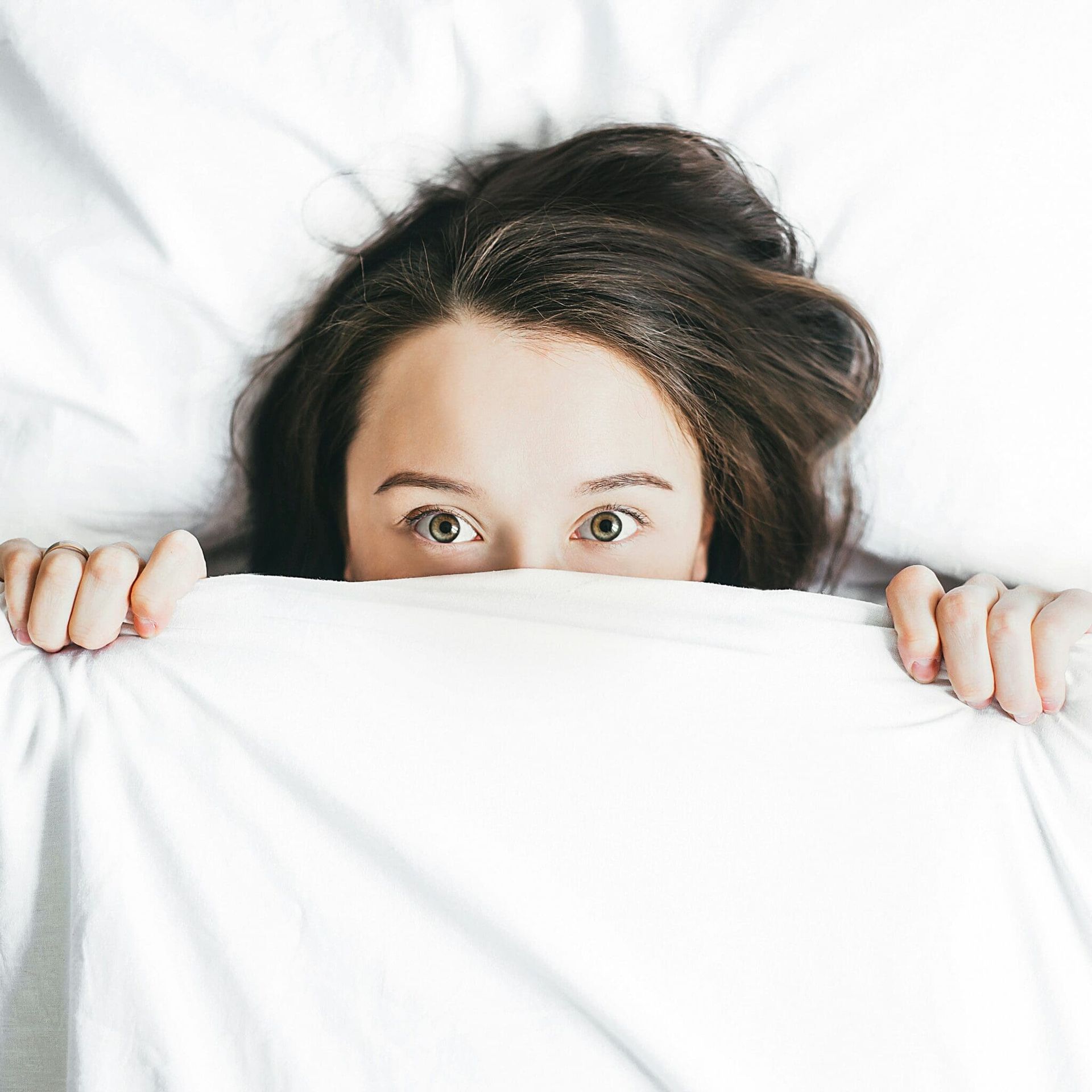 Woman peeking over a white blanket, eyes wide on a white bed