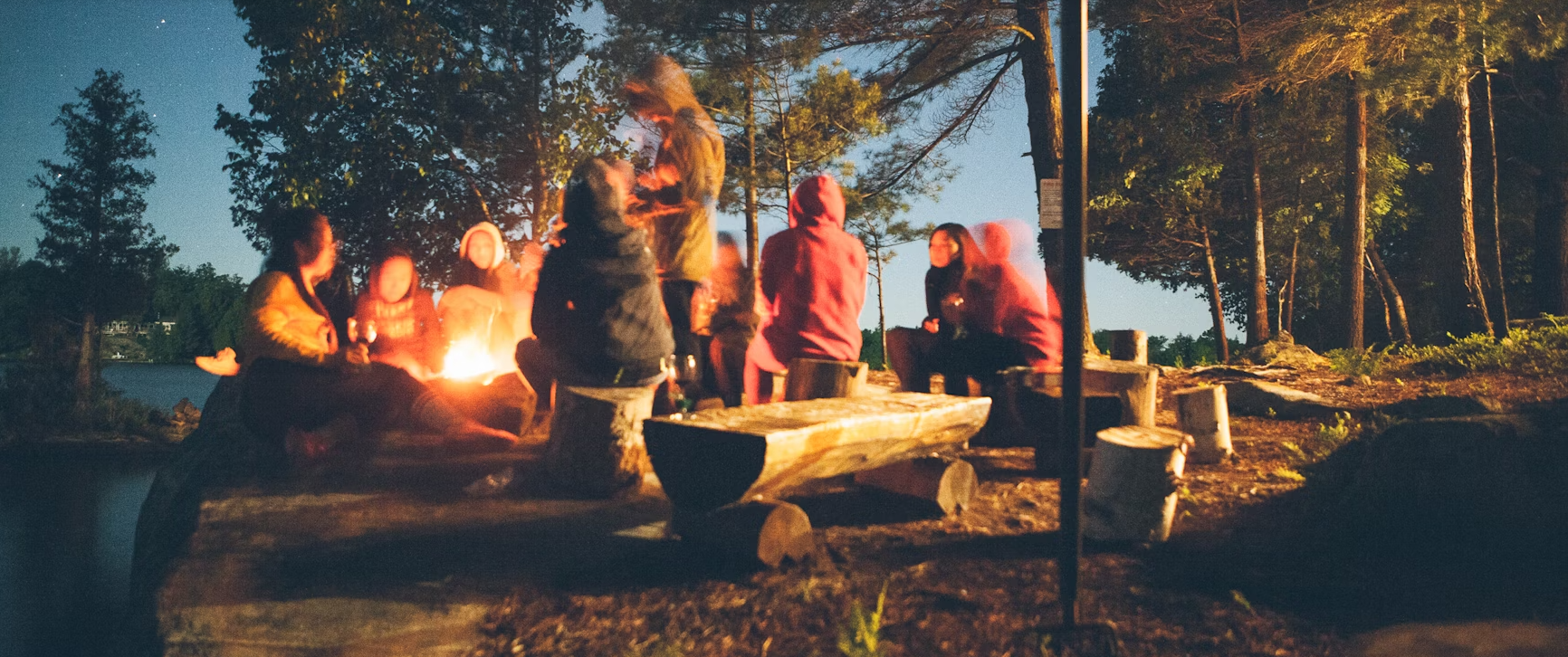 People gathered around a campfire outdoors at dusk, with glowing flames and trees in the background.