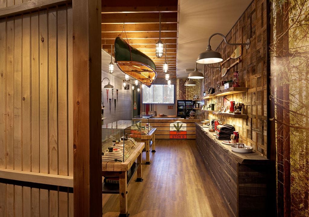 Warmly lit rustic café interior with wooden walls, hanging lights, shelves of bottles, and a long counter.