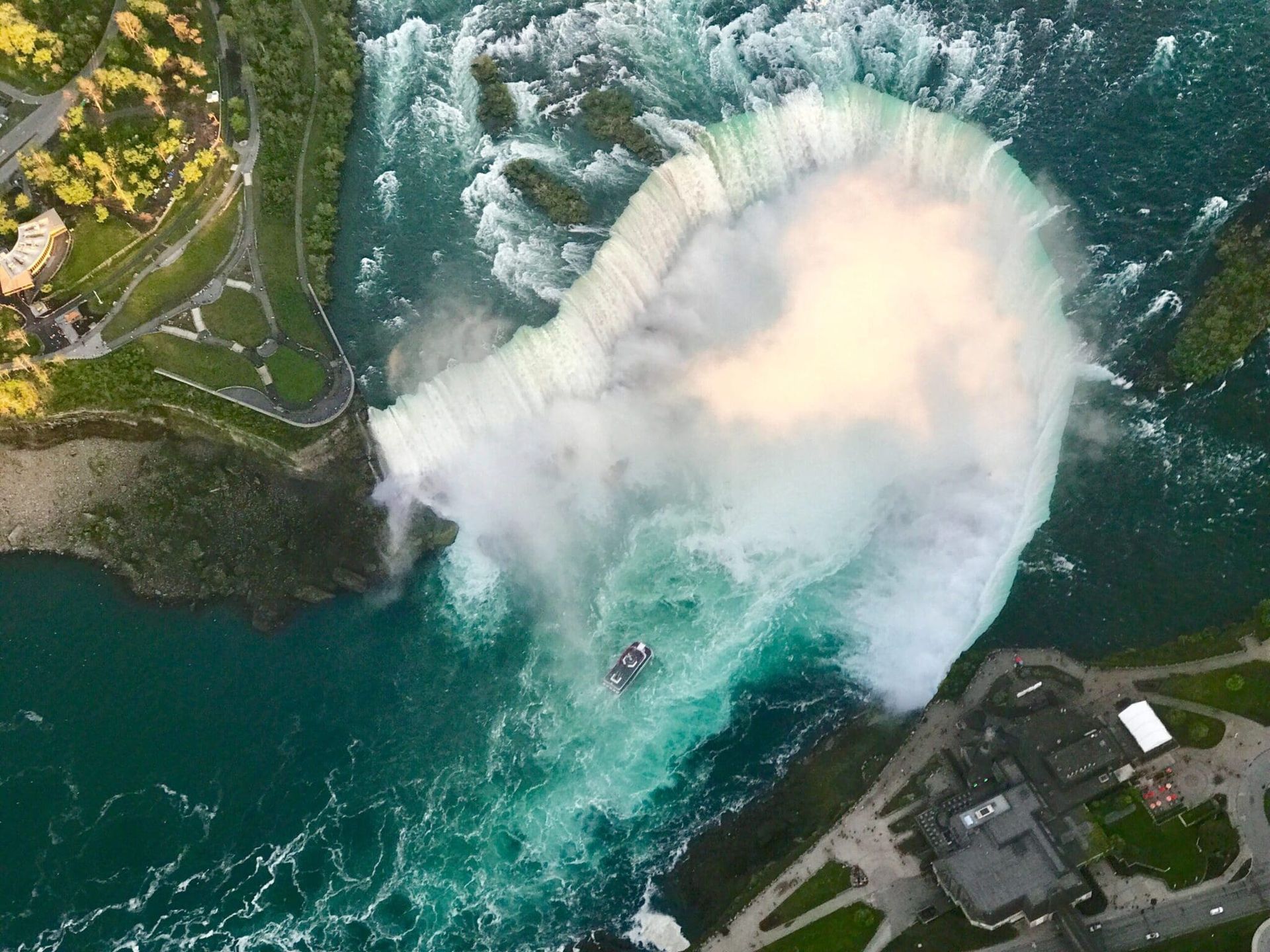 Aerial view of a large waterfall with white mist plunging into blue-green water near buildings and trees