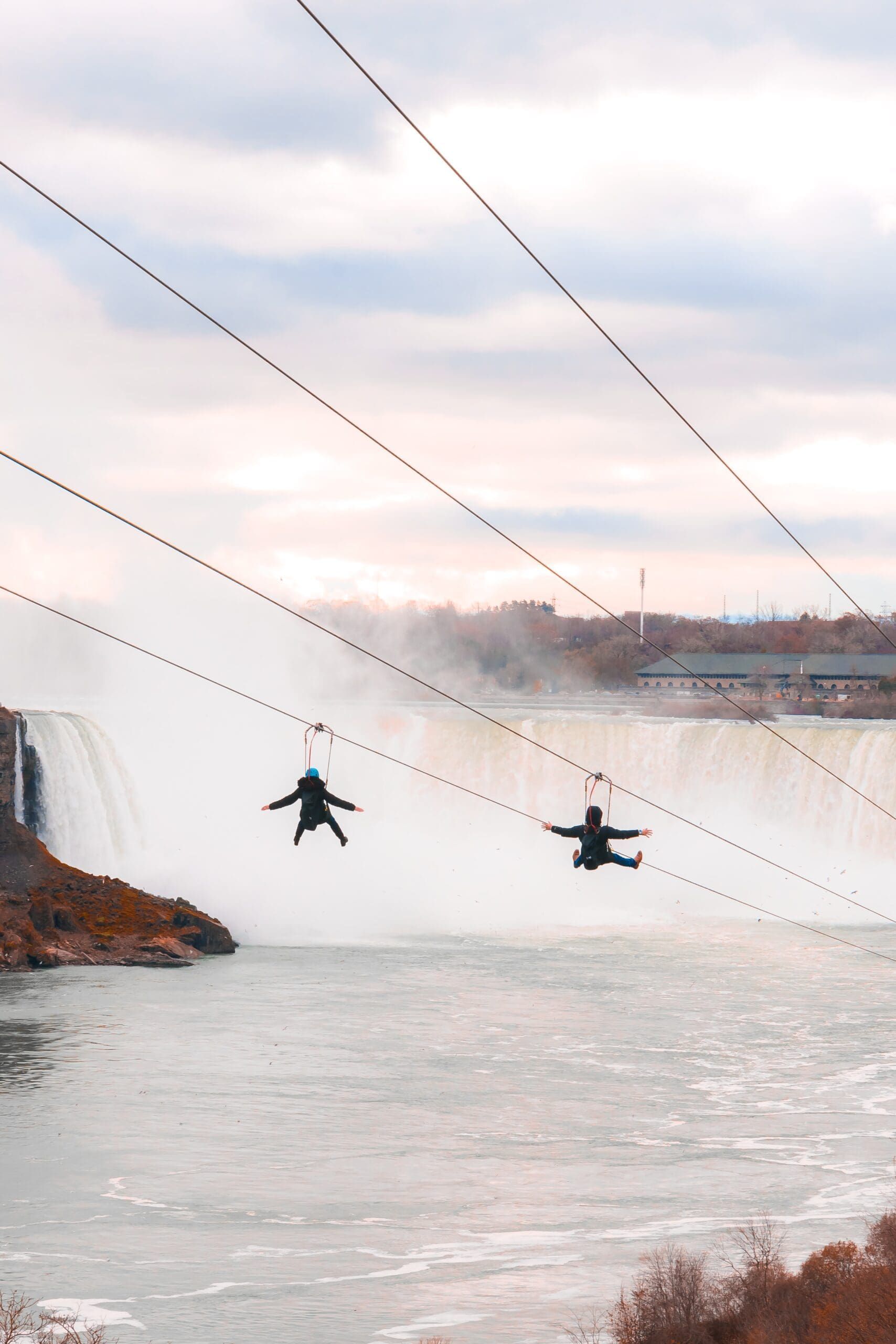Two people zip-lining over a wide river beneath a cloudy sky