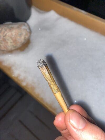 Hand holding a lit rolled cigarette with ash, near a countertop and a round stone object.