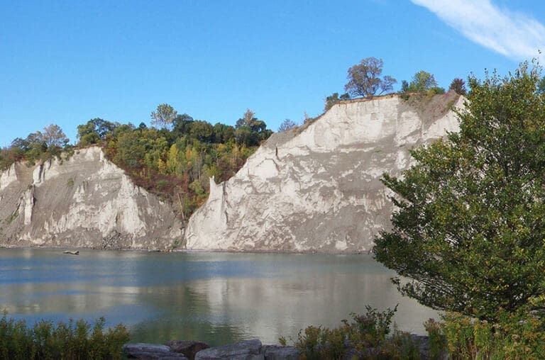 White chalk cliffs beside a calm lake under a blue sky, with trees along the shoreline