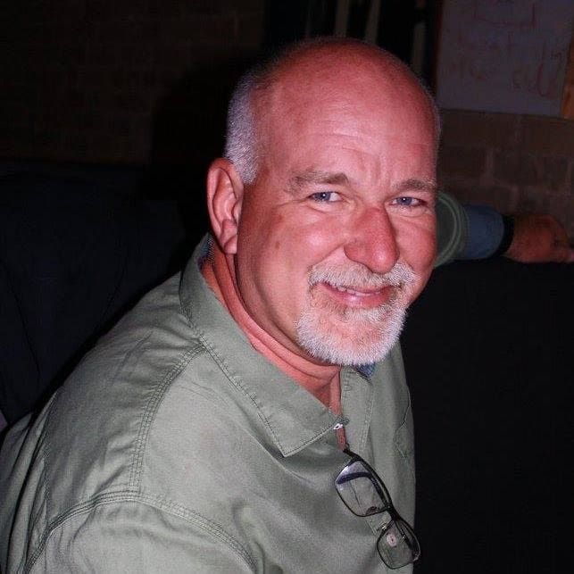 Smiling bald man in a green shirt, seated indoors with glasses hanging from his collar