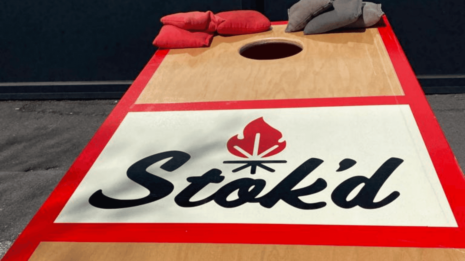 Cornhole board with red border and white center, featuring the word “Stoked” and two beanbags on top
