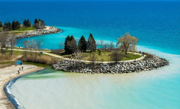 Small stone peninsula with trees in clear turquoise water and a sandy shore under a blue sky