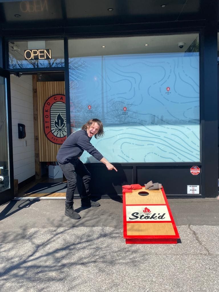 Person outdoors setting up a red cornhole board in front of a shop with an “OPEN” sign.