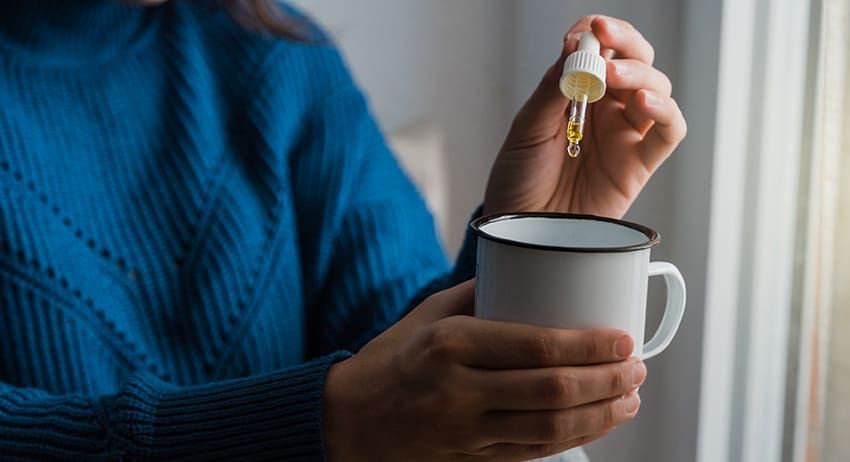 Hand holding a dropper above a white mug, adding liquid into a hot drink by a window.