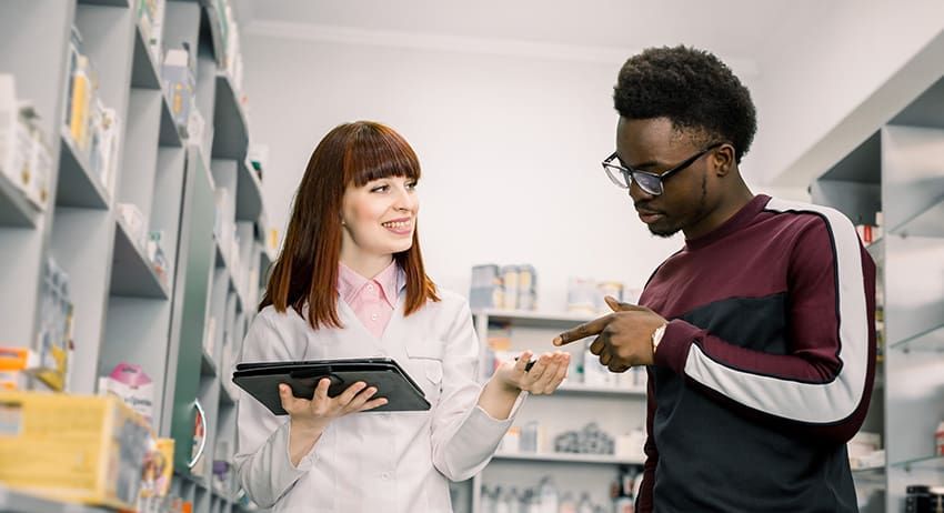 Two people in a pharmacy aisle discussing a tablet, with shelves of products in the background