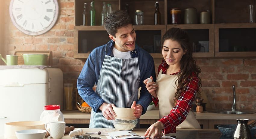 Couple cooking together in a rustic kitchen, smiling over a mixing bowl and countertop ingredients.