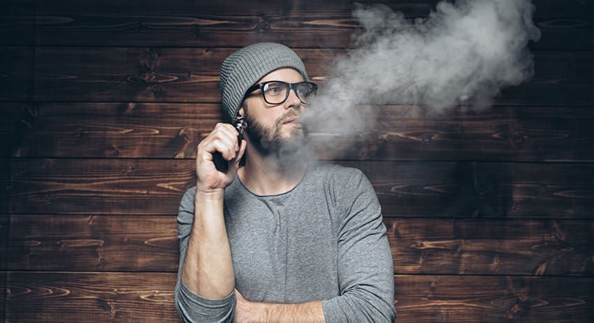 Person in gray shirt and beanie exhales vapor while holding a vape against a wooden wall.